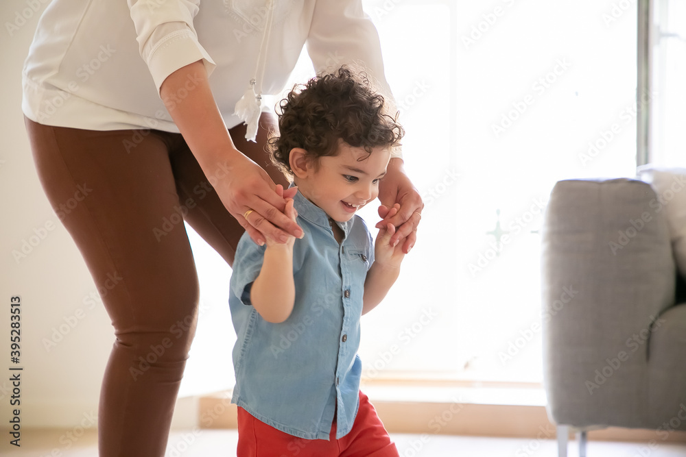 Fototapeta premium Funny mix-raced boy learning walking with help of mother, looking down and smiling. Cropped mother holding son hands and helping toddler in blue shirt. Family time, childhood and first step concept