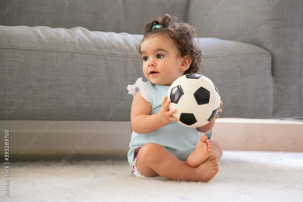 Positive black haired baby girl in pale blue clothes sitting on floor at home, looking away, playing soccer ball. Copy space. Kid at home and childhood concept Positive black haired baby girl in pale blue clothes sitting on floor at home, looking away, playing soccer ball. Copy space. Kid at home and childhood concept