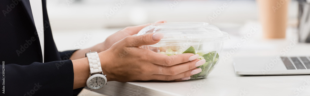 Naklejka premium Cropped view of businesswoman holding plastic bowl with food at work on blurred background, 