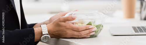 Cropped view of businesswoman holding plastic bowl with food at work on blurred background  