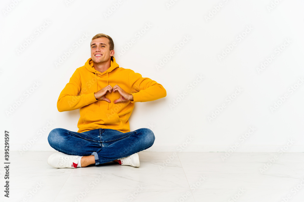 Young caucasian man sitting on the floor smiling and showing a heart shape with hands.