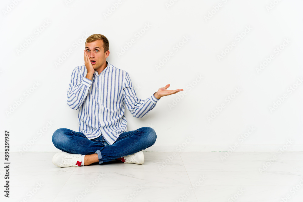 Young caucasian man sitting on the floor impressed holding copy space on palm.