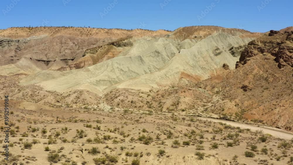 Dirt road winding in front of the large geologic formations and mountains in the Rainbow Basin in the Mojave Desert; drone pullback view. Dirt road winding in front of the large geologic formations and mountains in the Rainbow Basin in the Mojave Desert; drone pullback view.