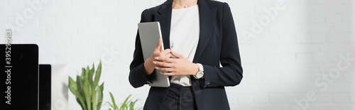 Cropped view of businesswoman in formal wear holding laptop near plants and computer monitors, 