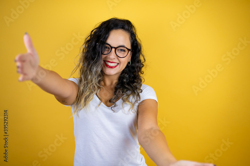 Young beautiful woman wearing casual white t-shirt over isolated yellow background looking at the camera smiling with open arms for hug