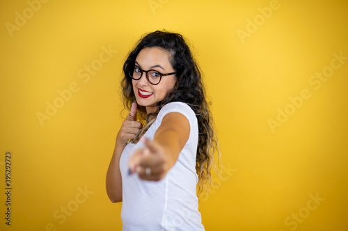 Young beautiful woman wearing casual white t-shirt over isolated yellow background pointing to you and the camera with fingers, smiling positive and cheerful