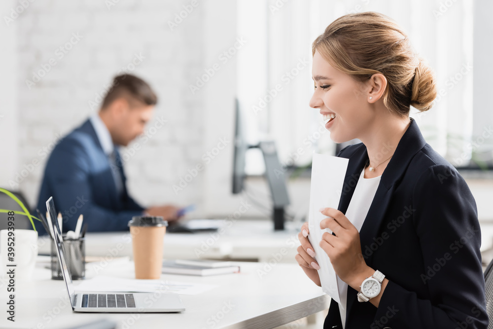 businesswoman with paper sheet looking at laptop, while sitting at workplace on blurred background
