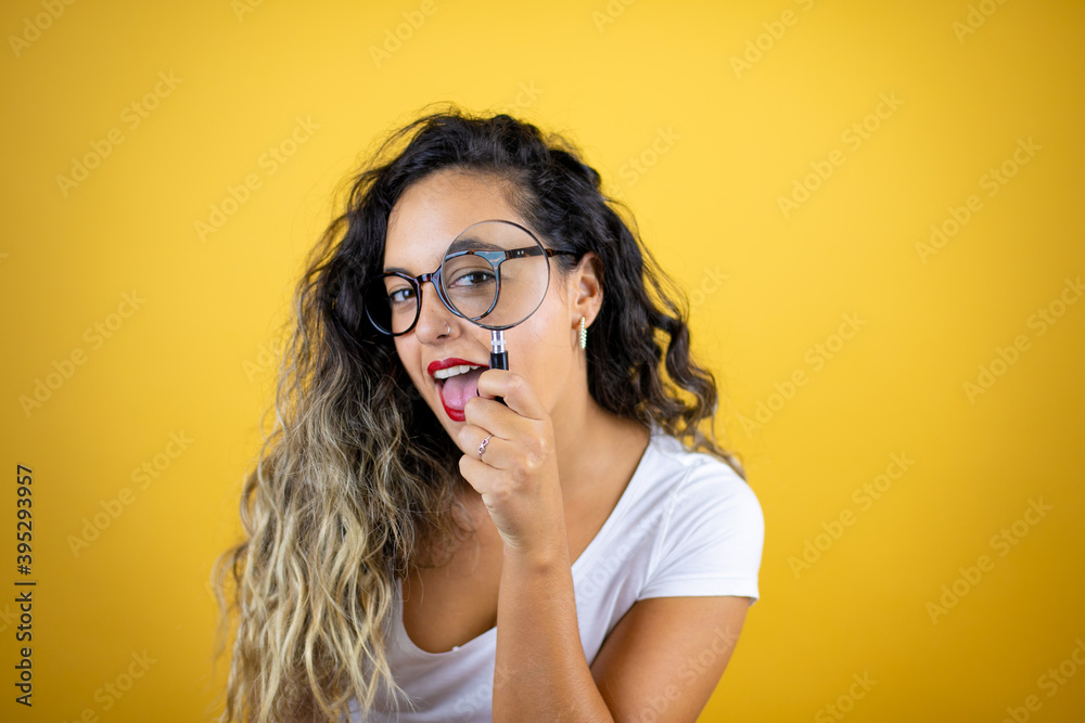Young beautiful woman wearing casual white t-shirt over isolated yellow background surprised looking through a magnifying glass