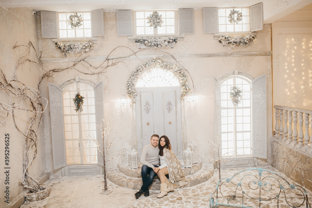 Adorable couple sit on stairs at the background Christmas decoration, cuddling, beautiful wife with loving husband spend winter holidays together, New Year celebration concept