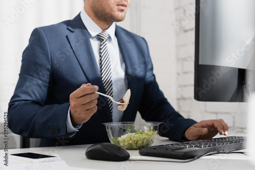 Cropped of businessman eating meal from plastic bowl, while typing on computer keyboard at workplace