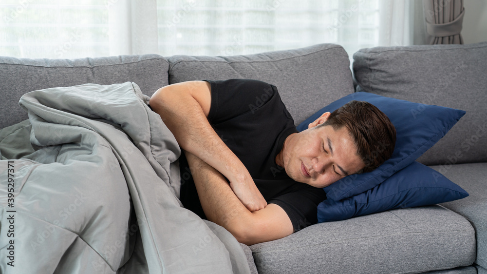 Sick and unwell Asian men holding his head, having a strong headache and sleeping on sofa at home wrapped in grey blanket Sick and unwell Asian men holding his head, having a strong headache and sleeping on sofa at home wrapped in grey blanket