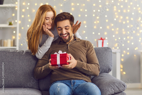 Murais de parede Joyful excited man sits on the sofa in the room and receives a gift from his girlfriend