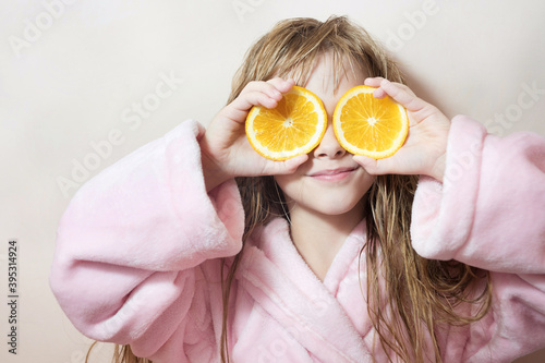 charming blonde girl with wet hair in a pink bath robe covers her eyes with oranges on a pink background
