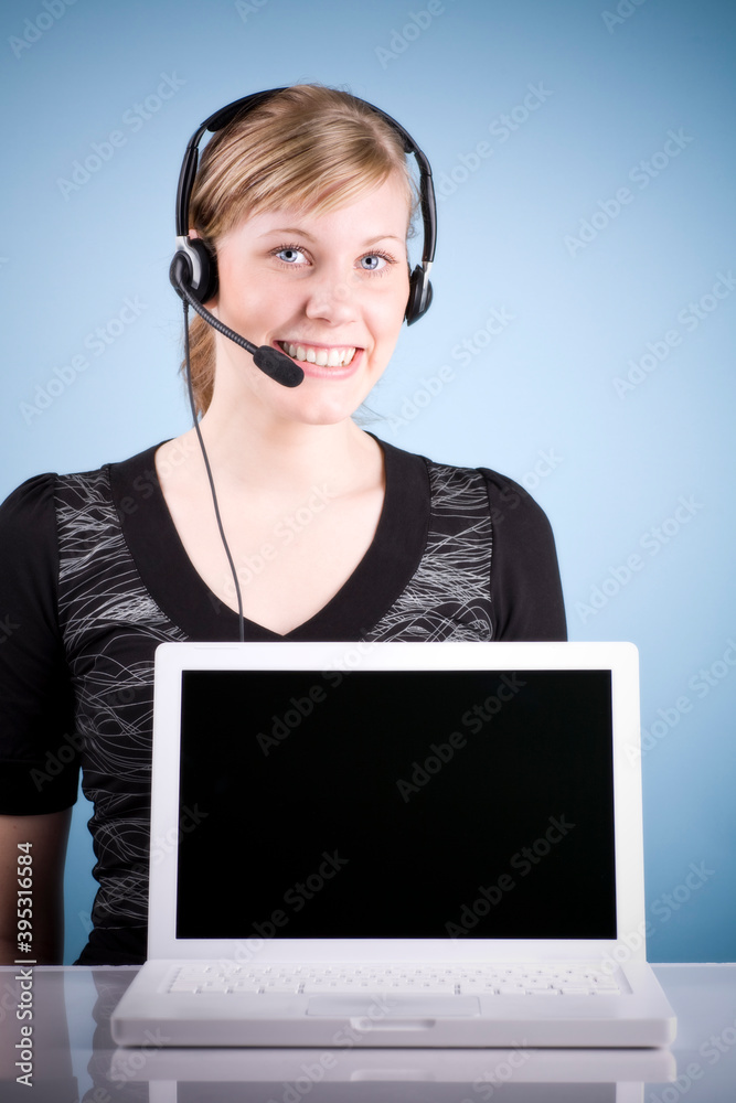 Blonde smiling girl with headset in front of laptop. Studio photo isolated on light blue background. Blonde smiling girl with headset in front of laptop. Studio photo isolated on light blue background.