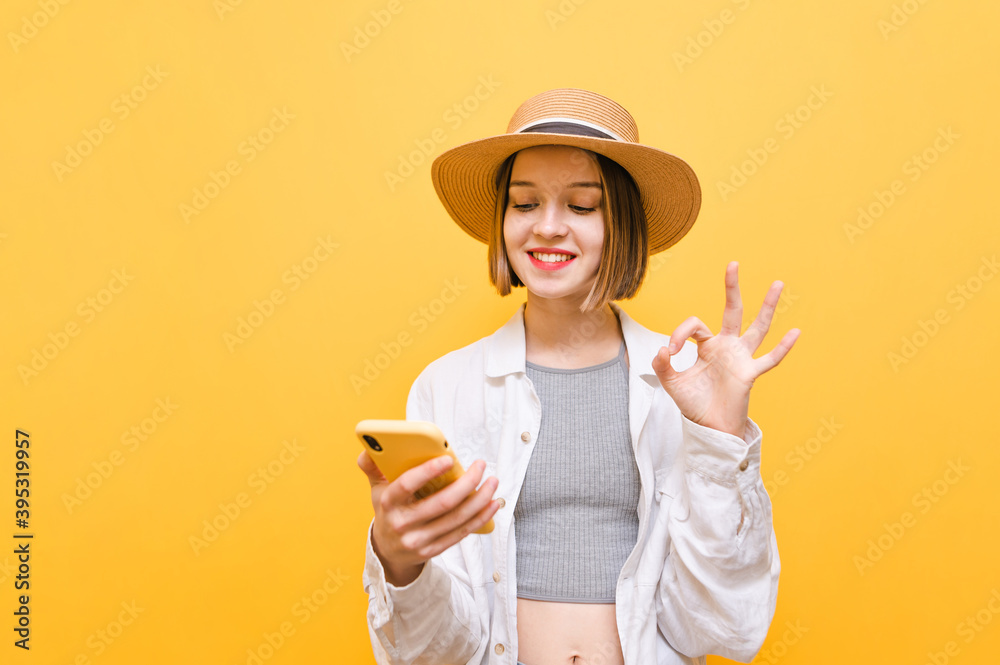 Smiling girl uses smartphone on yellow background and shows OK gesture, wears summer light clothes and hat. Positive lady looks at smartphone screen with smile on her face and shows gesture Smiling girl uses smartphone on yellow background and shows OK gesture, wears summer light clothes and hat. Positive lady looks at smartphone screen with smile on her face and shows gesture