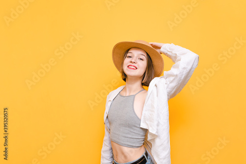 Smiling lady in summer dress and hat poses against yellow background with happy face, looks into camera and wears hat and shirt. Cheerful lady tourist isolated on orange background. Copy space