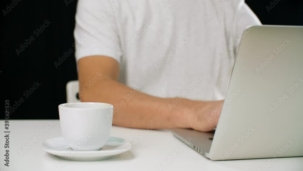 Close-up view of man using laptop and holding white cup. Cropped shirt of man in white t-shirt working on laptop computer and drinking coffee on black background Close-up view of man using laptop and holding white cup. Cropped shirt of man in white t-shirt working on laptop computer and drinking coffee on black background