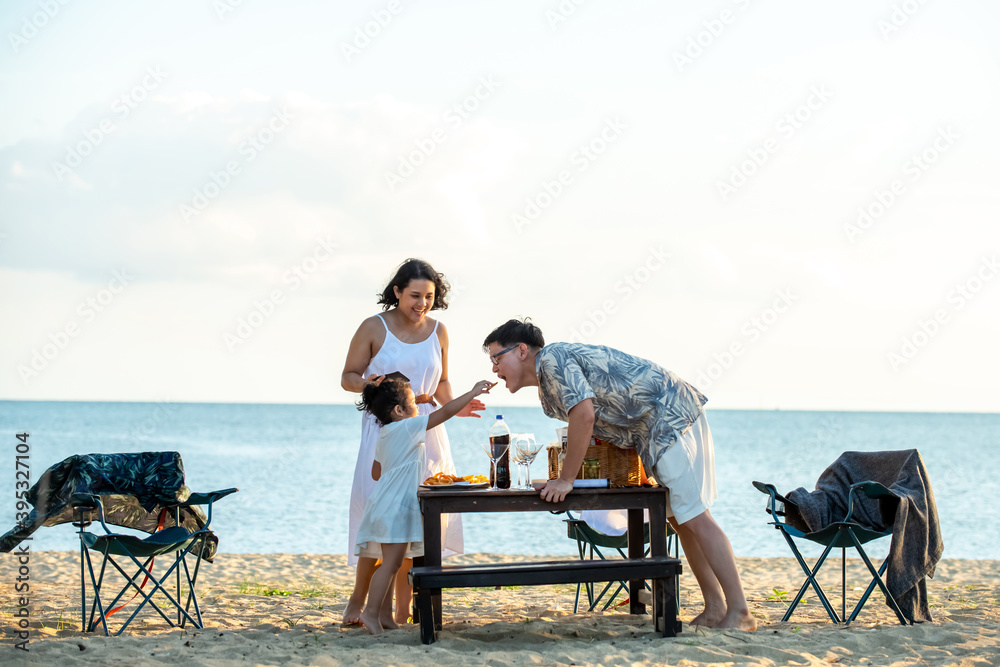 Asian couple man and woman with little daughter enjoy dinner party together on island beach at summer sunset. Happy family parents with cute child girl relax and having fun with on holiday vacation