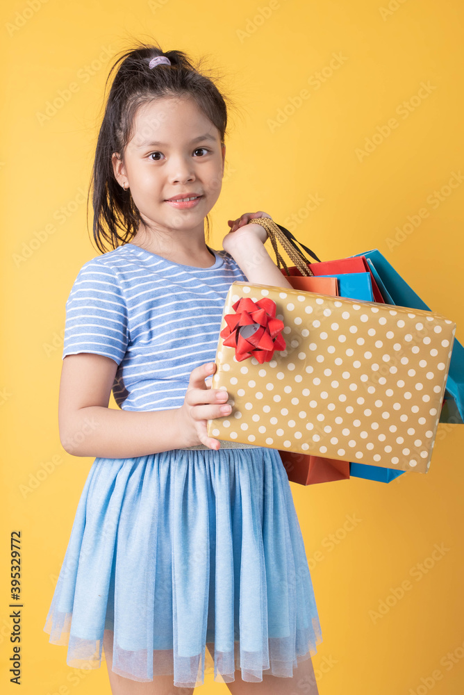 Portrait of little girl wear skirt holding shopping bags and while standing and looking at camera isolated over yellow background. happy child holding gift box with his family. Portrait of little girl wear skirt holding shopping bags and while standing and looking at camera isolated over yellow background. happy child holding gift box with his family.