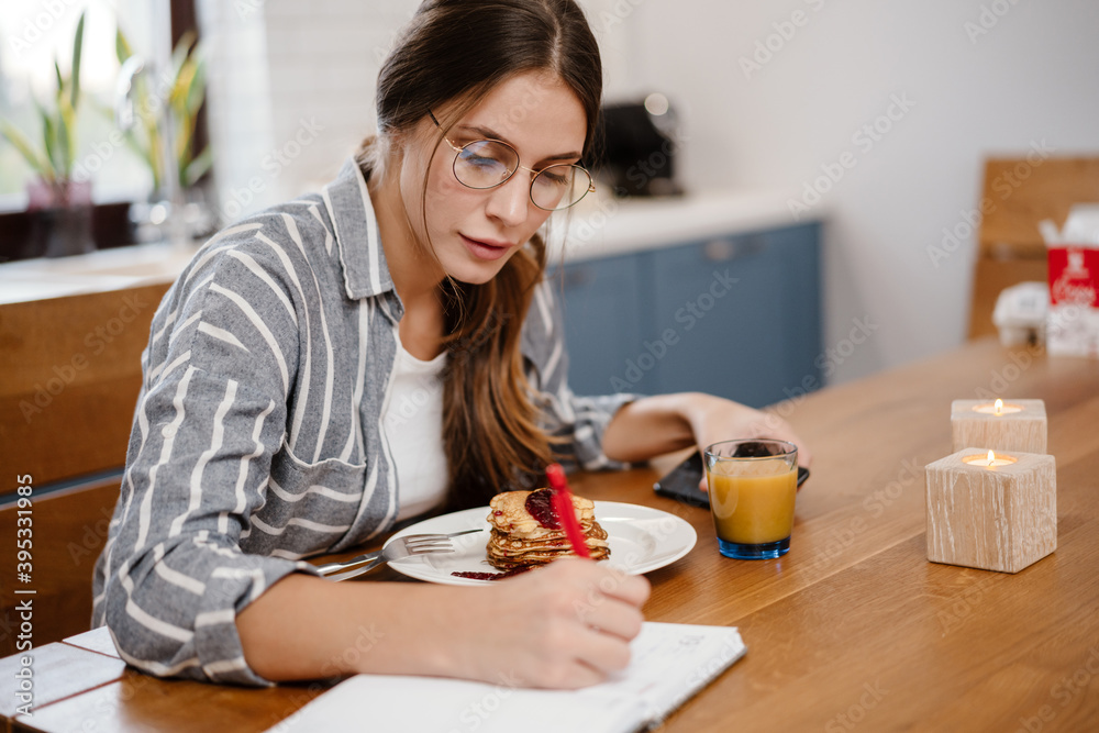 Woman writing down notes and using cellphone while having breakfast Woman writing down notes and using cellphone while having breakfast