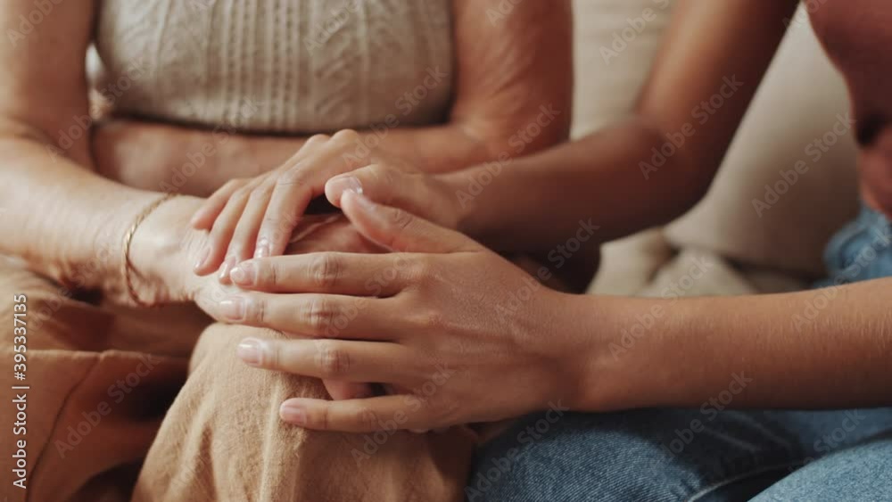 Close up selective focus shot of hands of young woman holding hands of elderly grandmother while sitting with her on sofa at home