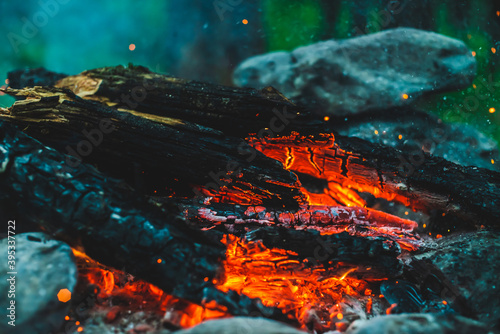 Vivid smoldered firewoods burned in fire close-up. Atmospheric warm background with orange flame of campfire and blue smoke. Full frame image of bonfire. Beautiful whirlwind of embers and ashes in air