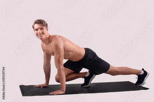Smiling shirtless man with muscular body doing yoga or pilates on sports mat over light studio background