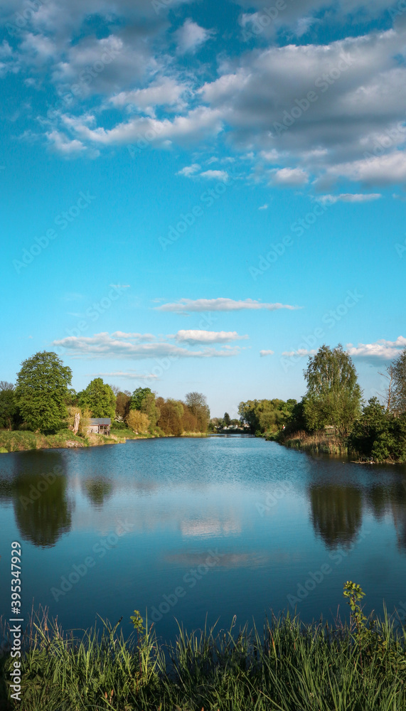 A large pond in the village. Blue water and sky. Greens around.
