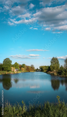 A large pond in the village. Blue water and sky. Greens around. © Olha