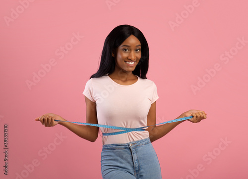 Perfect size. Slim black lady measuring her waist, happy with slimming diet over pink studio background © Prostock-studio