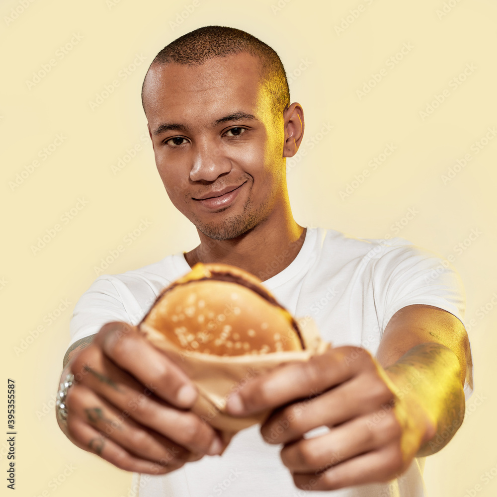 Eating food. A dark-skinned male with tattoos holding a burger in hands while smiling and looking into a camera Eating food. A dark-skinned male with tattoos holding a burger in hands while smiling and looking into a camera