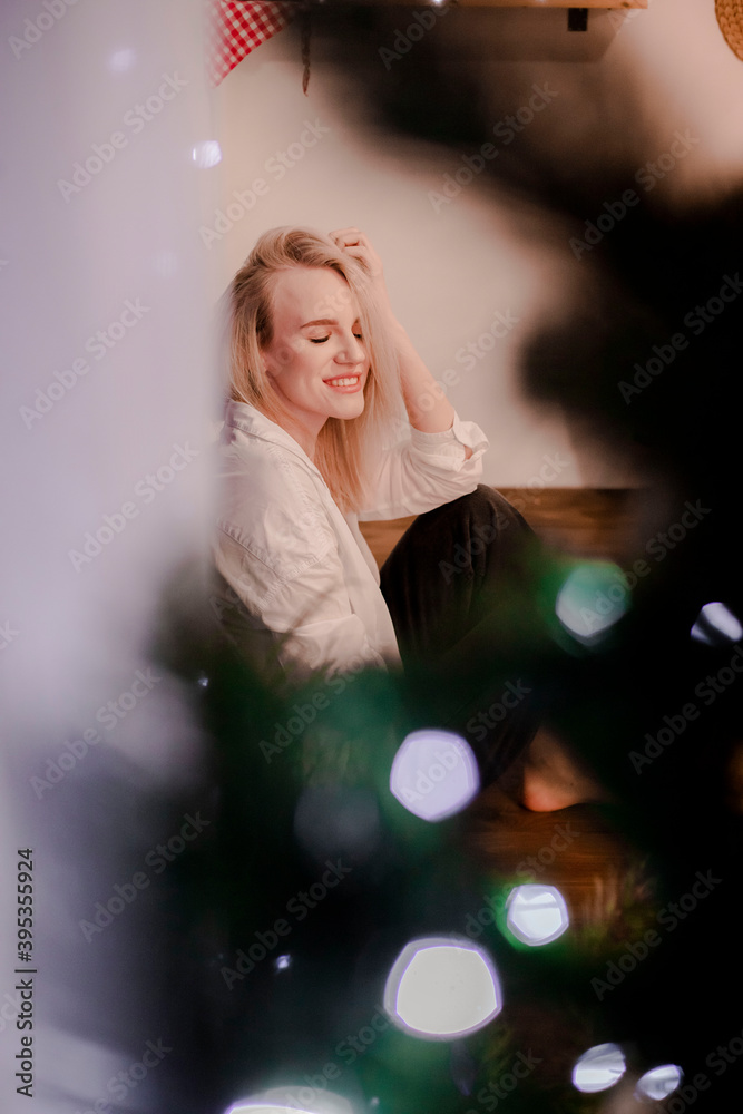 A nice, young blonde girl alone in a white shirt and jeans in the 
kitchen. The kitchen is decorated with New Year's decor. New Years 
is soon.