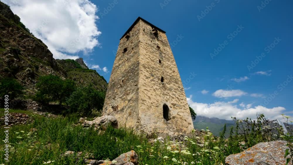 An ancient tower to protect against enemies in the mountains of the north Caucasus. Around the tower destroyed the old city. Timelaps. Floating clouds in the blue sky. Low angle An ancient tower to protect against enemies in the mountains of the north Caucasus. Around the tower destroyed the old city. Timelaps. Floating clouds in the blue sky. Low angle