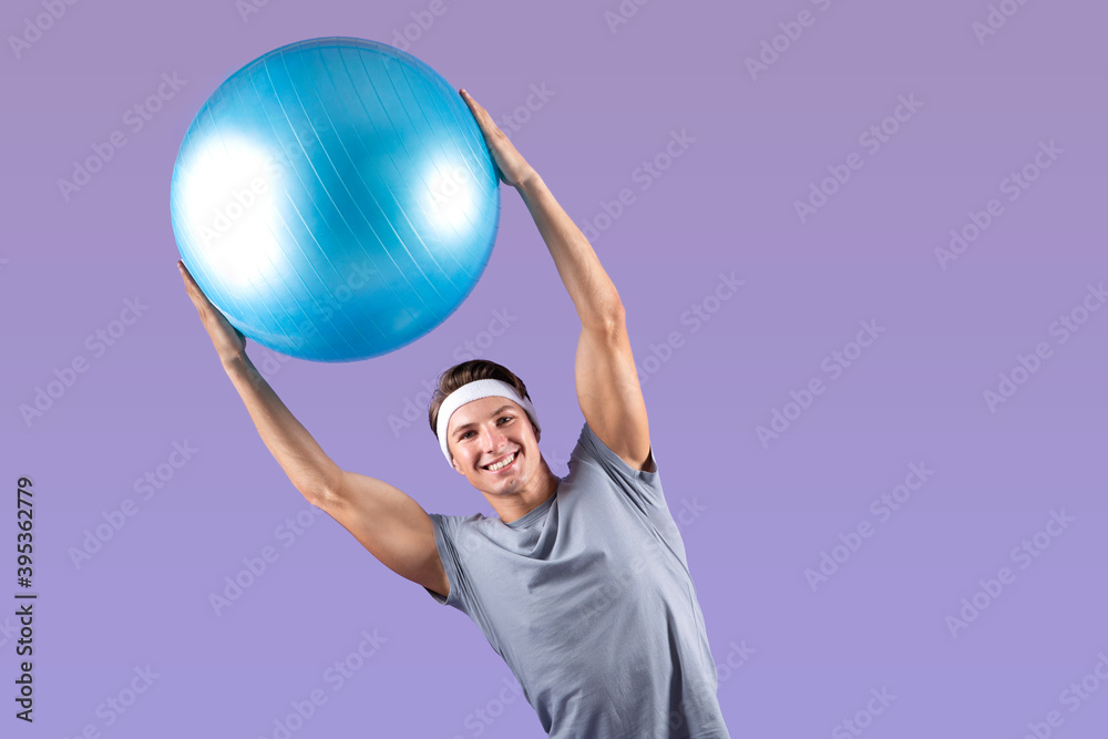 Positive young sportsman lifting fitness ball above his head, bending aside on lilac studio background, copy space Positive young sportsman lifting fitness ball above his head, bending aside on lilac studio background, copy space