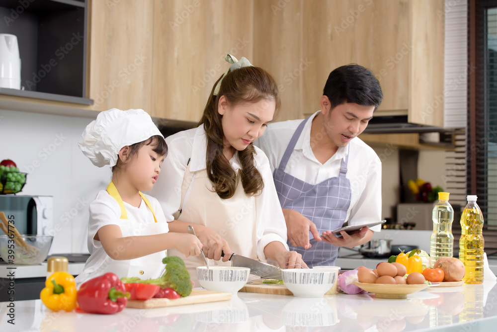 Asian parents are teaching their daughters to easy home cook while looking at how to cook on a tablet. Family enjoys cooking together home cook in kitchen at home. Focusing on Mom center. Asian parents are teaching their daughters to easy home cook while looking at how to cook on a tablet. Family enjoys cooking together home cook in kitchen at home. Focusing on Mom center.