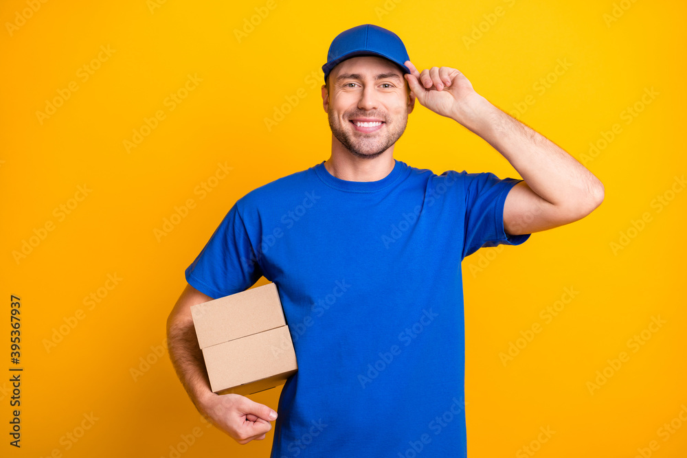 Portrait of cheerful working man arm touch cap hold carton package isolated on yellow color background