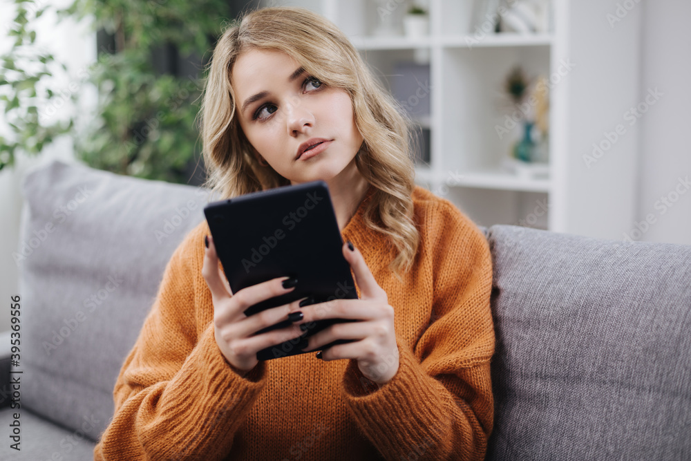 Dreamy young blonde in brown sweater relaxing on couch and holding digital tablet. Pretty woman using modern gadget during free time at home. Dreamy young blonde in brown sweater relaxing on couch and holding digital tablet. Pretty woman using modern gadget during free time at home.