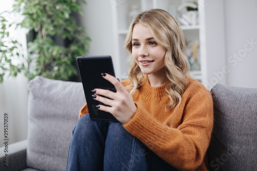 Positive young woman in casual clothing using digital tablet for video conversation while staying at home. Concept of communication on distance.