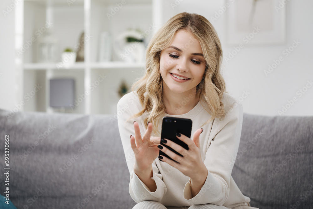 Charming young lady in white domestic clothing sitting on cozy sofa and browsing internet on modern smartphone. Happy woman with blond hair enjoying free time at home. Charming young lady in white domestic clothing sitting on cozy sofa and browsing internet on modern smartphone. Happy woman with blond hair enjoying free time at home.