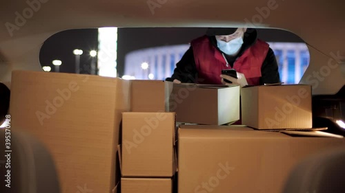 Delivery service man wearing his protective mask due to Covid-19 late nigt with a delivery item in a car. Volunteer Work in the Midst of an Epidemic. Courier with a Cardboard Box Package on a second