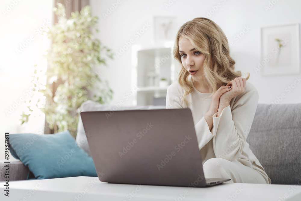 Pretty young female in domestic clothing sitting on sofa and talking during video call. Attractive woman with blond hair using laptop for conversation. Pretty young female in domestic clothing sitting on sofa and talking during video call. Attractive woman with blond hair using laptop for conversation.