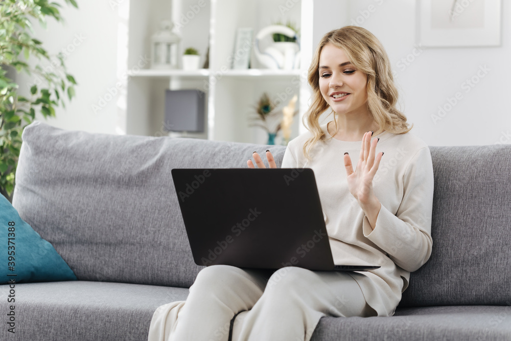 Fototapeta premium tractive young blonde dressed in white clothing smiling and gesturing during video chat. Cheerful woman holding opened laptop on knees while sitting on cozy sofa.