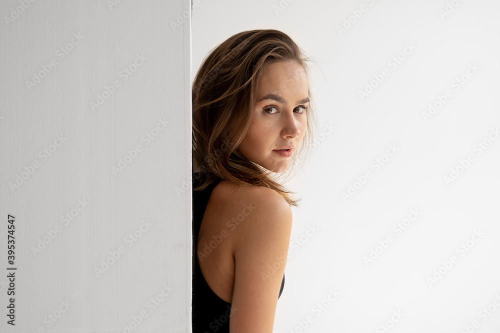 Portrait of young brunette skinny woman is posing in black underwear and standing behind a white wall in white studio. Model tests of pretty girl in basic clothes on cyclorama. Portrait of young brunette skinny woman is posing in black underwear and standing behind a white wall in white studio. Model tests of pretty girl in basic clothes on cyclorama.