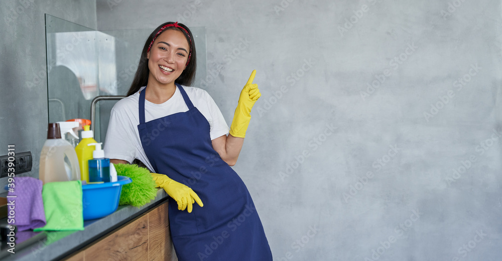 Cheerful young woman, cleaning lady in protective gloves smiling at camera, pointing up while standing in the kitchen with cleaning products and equipment, ready for cleaning the house Cheerful young woman, cleaning lady in protective gloves smiling at camera, pointing up while standing in the kitchen with cleaning products and equipment, ready for cleaning the house