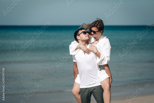 Young Caucasian man carrying his smiling girlfriend on his back while enjoying a late afternoon together at the beach. Phuket. Thailand.