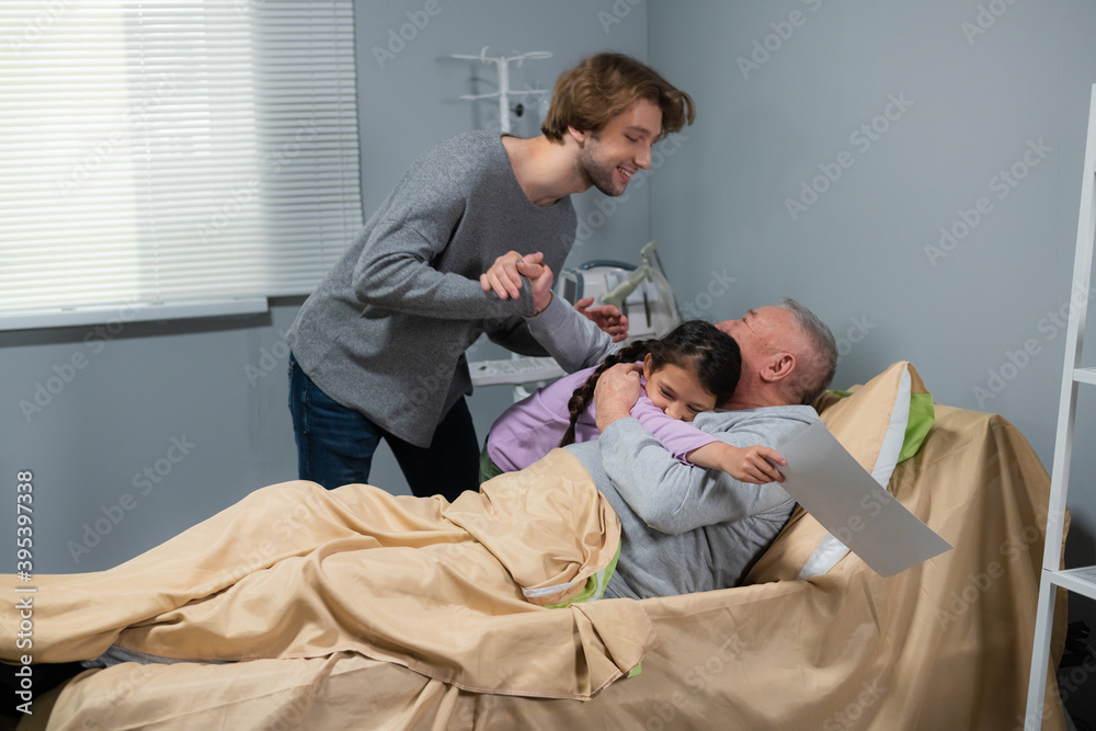 Obraz premium Grandchildren, a little girl and her elder brother, visiting their grandpa in a hospital ward, the little girl is hugging her grandpa and the older boy is shaking his hand.