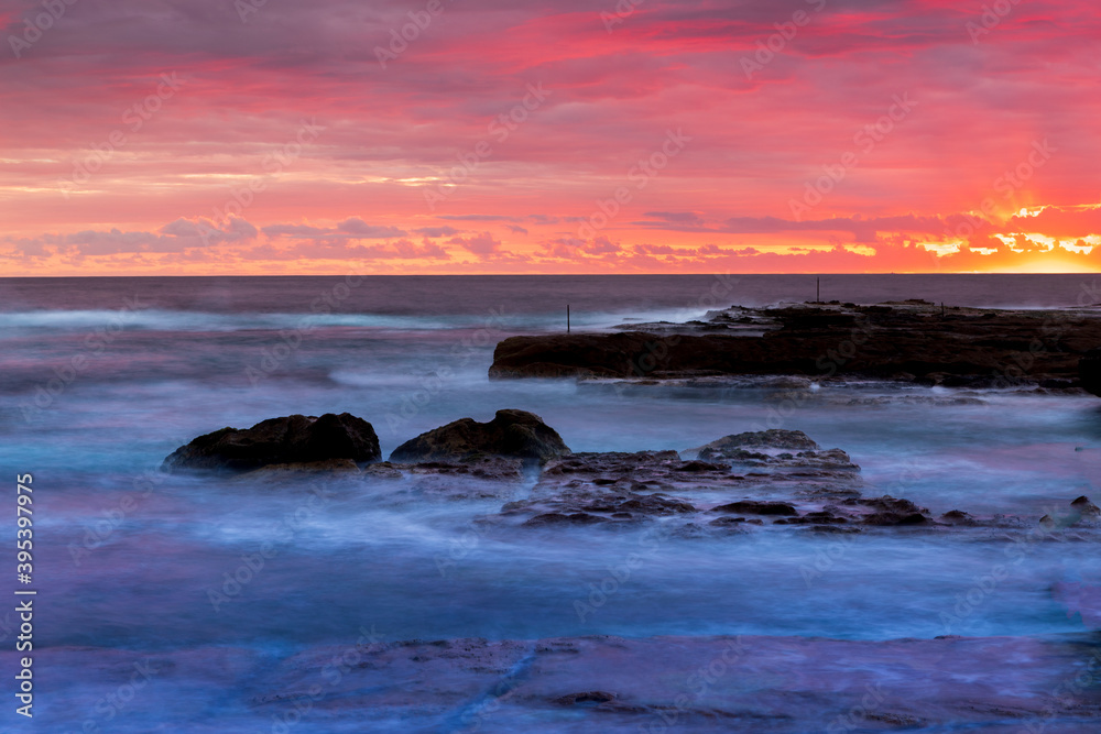 Obraz premium Rays of rising sun highlight clouds in the sky with red magenta colour. Calm ocean with rocks hit by waves is under beautiful sky