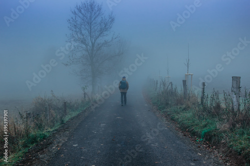 Man walking on scary misty road. Moody blue fog.Mystical fantasy Halloween atmosphere.Person walking to adventure.Horor like scenery.Dark moody landscape with pathway leading through.Mysterious park.