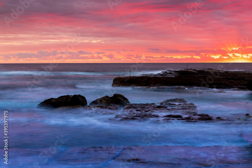 Rays of rising sun highlight clouds in the sky with red magenta colour. Calm ocean with rocks hit by waves is under beautiful sky