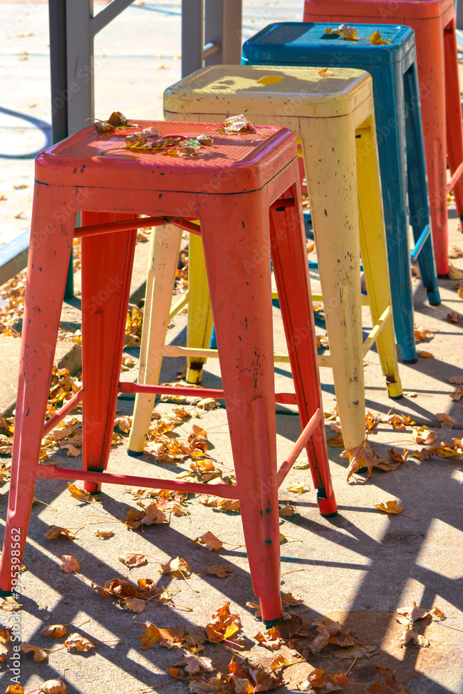Colorful metal stools outside a restaurant on a beautiful fall day with leaves decorating the scene. Red, yellow, orange and turquoise stools.